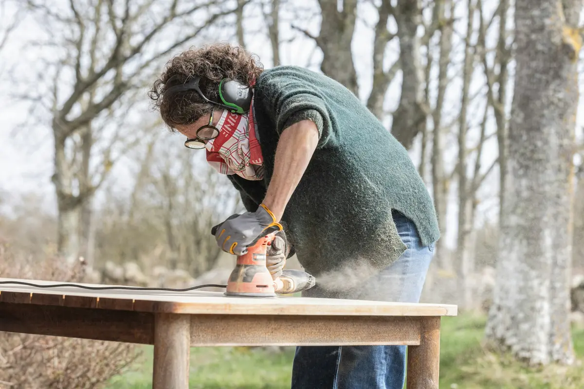 Une artisane ponce un plateau de table en extérieur.